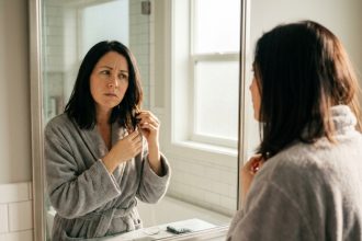 Femme aux cheveux bruns dans une salle de bain moderne qui observe sa chute de cheveux dans le miroir, exprimant une légère inquiétude
